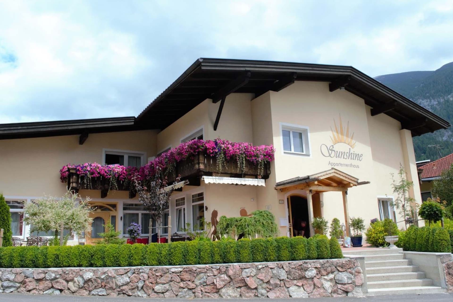 Das Appartementhaus Sunshine mit Sonnenlogo und Blumenbalkonen, dahinter die Berge