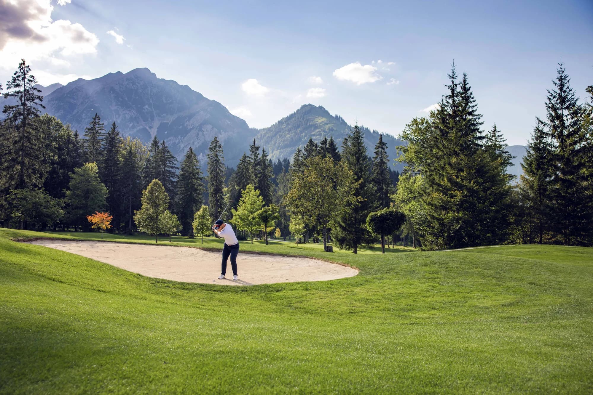 Golferin schlägt aus dem Bunker, ein Mitspieler schaut zu, im Hintergrund das Karwendel