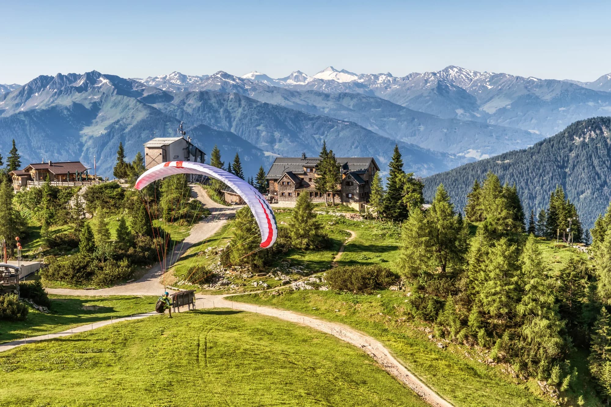 Gleitschirmpilot startet bei einer Berghütte, im Hintergrund die schneebedeckten Alpen