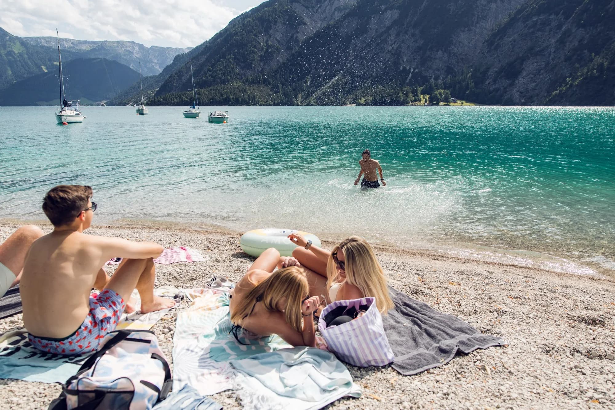 Freunde am Kiesstrand des Achensees, einer kommt aus dem türkisblauen Wasser, dahinter ankern Segelboote