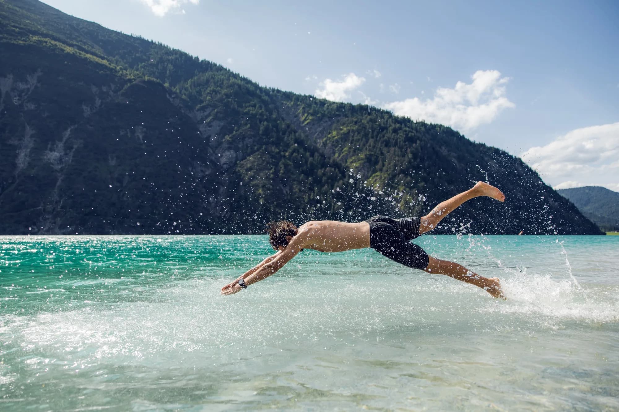 Junger Mann springt kopfüber in das türkisblaue Wasser des Achensees, dahinter bewaldete Berghänge